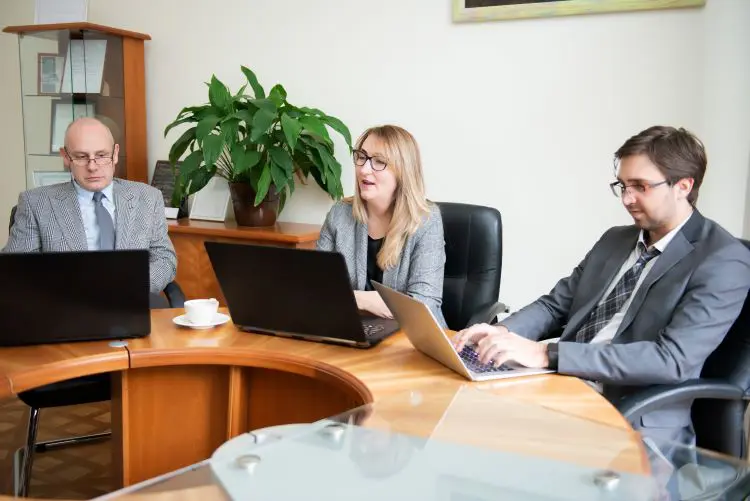 Three suited business people sit around a round conference table together looking and their laptops.
